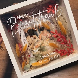 May include: A white framed shadow box with a wedding photo, dried flowers, and the words "Mein Brautstrau&szlig;" in white script. The photo is of a couple, with the names "Lea & Max" and the date "03. Oktober 2025" visible.