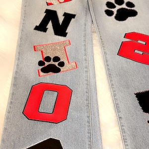 May include: Light blue denim jeans with red, black, and silver glitter patches spelling out "SENIOR" on the left leg and "PANTHERS" and the number "2" on the right leg.