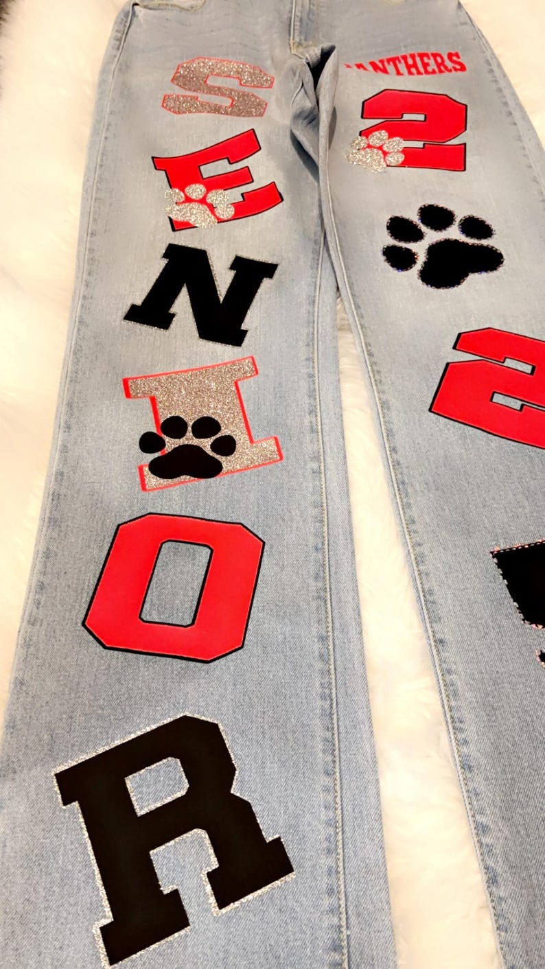 May include: Light blue denim jeans with red, black, and silver glitter patches spelling out "SENIOR" on the left leg and "PANTHERS" and the number "2" on the right leg.
