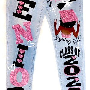 May include: Light blue denim jeans with pink glitter and black lettering that says "Senior" on the left leg and "Signing Out Class of 2025" on the right leg. The right leg also features a black silhouette of a woman wearing a graduation cap and gown.