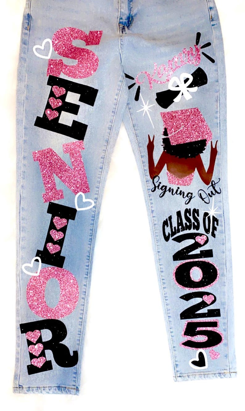 May include: Light blue denim jeans with pink glitter and black lettering that says "Senior" on the left leg and "Signing Out Class of 2025" on the right leg. The right leg also features a black silhouette of a woman wearing a graduation cap and gown.