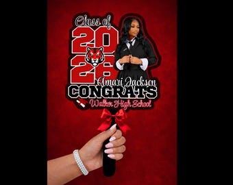 May include: A graduation sign with the text "Class of 2023" and "Congrats Amari Jackson Walker High School". The sign features a photo of a graduate and a red bow. The sign is held by a hand wearing a bracelet.