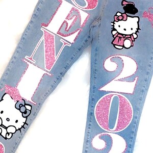 May include: A pair of blue denim jeans with pink glitter lettering that says "SENI 2025" and a pink glitter Hello Kitty graphic with a graduation cap.