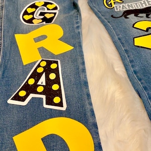 May include: Blue denim jeans with yellow and black fabric appliques spelling out "GRAD" and "8th GRADE". The jeans also have a black and yellow applique of a graduation cap, a black and yellow applique of a panther, and a black and yellow applique of a shoe.