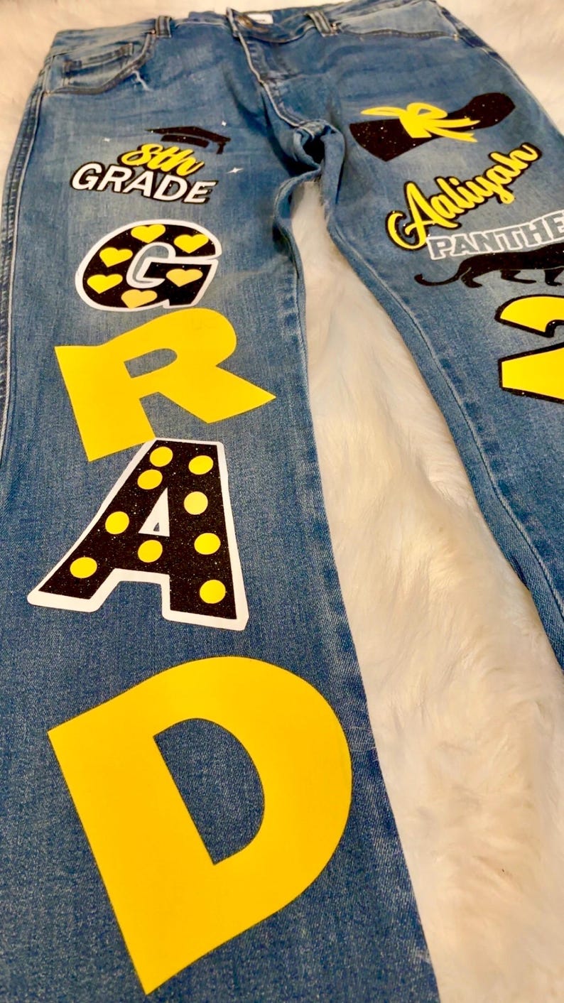 May include: Blue denim jeans with yellow and black fabric appliques spelling out "GRAD" and "8th GRADE". The jeans also have a black and yellow applique of a graduation cap, a black and yellow applique of a panther, and a black and yellow applique of a shoe.
