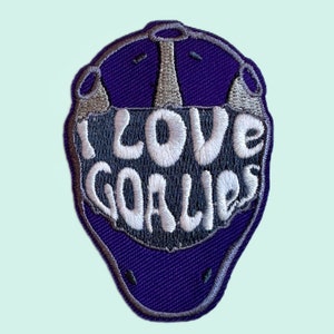 以下が含まれることがあります： 紫とグレーの刺繍パッチで、白い文字で「I LOVE GOALIES」と書かれています。