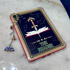 May include: A phone case with a gold border and a design featuring a sword through an open book, with the text "THE READER". A charm with a heart, stars, and a moth is attached.