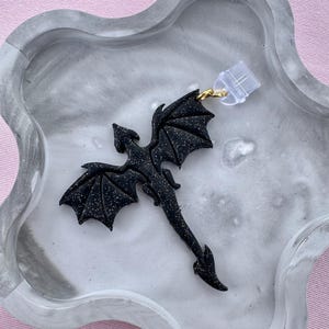 Könnte beinhalten: Ein schwarzer Drachenanhänger mit Glitzerakzenten, befestigt an einem transparenten Kunststoffverschluss mit einem goldfarbenen Metallring. Der Drache liegt auf einem grau-weißen Marmortablett vor rosa Hintergrund.