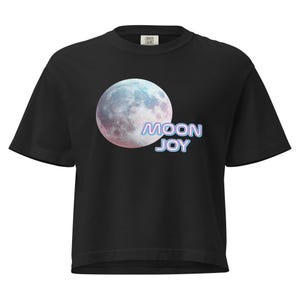 May include: Black t-shirt featuring a large, colorful moon graphic with pink, blue, and white hues. The text "MOON JOY" is printed below the moon in a gradient blue and purple font. The shirt is a crew neck style.
