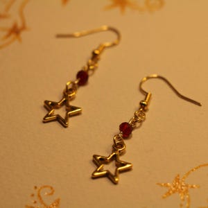 Könnte beinhalten: Ein Paar goldfarbene Ohrringe in Sternform mit roten Perlen. Die Ohrringe haben einen Hakenverschluss. Die Sternanhänger sind hohl und haben ein fünfzackiges Design. Sie liegen auf einer hellen Oberfläche.