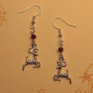 Puede incluir: Un par de pendientes colgantes plateados con un dije de reno y una pequeña cuenta roja. Los pendientes cuelgan de un gancho plateado. El fondo es de color beige claro con detalles de estrellas doradas brillantes.