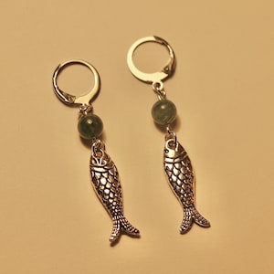 Puede incluir: Un par de pendientes de plata con forma de pez con cuentas de piedras preciosas verdes. Los pendientes tienen un diseño simple de aro y cuelgan del lóbulo de la oreja.