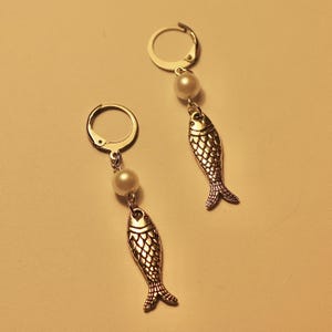 May include: A pair of silver fish earrings with white pearl accents. The earrings are dangle style with a hoop closure.