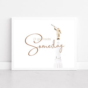 May include: A white framed print with a brown inscription that reads "I'll go inside someday". The inscription is in a cursive font. The print features a black and white photograph of a tall, white building with a golden statue of an angel on top. The angel is holding a trumpet.