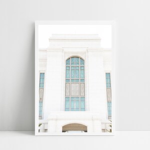May include: A framed print depicting a white building with a large arched window. The building features a classical design with columns and a detailed facade. The window has a grid pattern with blue-tinted glass. The print is displayed on a white surface.
