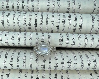 May include: A silver ring featuring a moonstone gemstone. The ring is displayed on a stack of books with printed text. The moonstone has an iridescent blue and white colour.