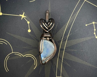 May include: A silver wire wrapped pendant featuring a crescent-shaped moonstone with a blue iridescent sheen. The pendant is set against a black background with a gold constellation design.