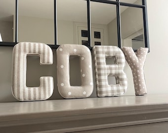 May include: Four fabric-covered letters spelling the name COBY. The letter C features beige and white vertical stripes, the O has a beige background with white polka dots, the B has a beige and white gingham check, and the Y has beige stars.