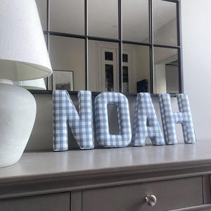 Könnte beinhalten: Der Name "NOAH" ist in großen, freistehenden Buchstaben geschrieben. Jeder Buchstabe ist mit einem blau-weiß karierten Stoff bedeckt. Die Buchstaben werden auf einer grauen Kommode mit einem Spiegel im Hintergrund angezeigt.