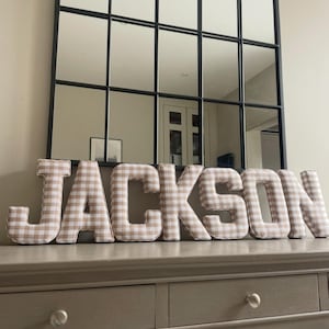 Könnte beinhalten: Stoffbuchstaben mit der Aufschrift "JACKSON" in einem beige-weißen Vichy-Muster. Die Buchstaben werden auf einer grauen Kommode angezeigt, mit einem großen Spiegel mit schwarzem Rahmen im Hintergrund.