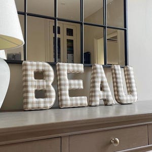 May include: The word "BEAU" spelled out in large, padded letters. Each letter is covered in a beige and white gingham fabric. The letters are displayed on a gray surface, with a mirror and lamp in the background.