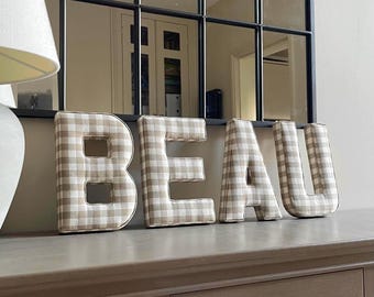 May include: The word "BEAU" spelled out in large, padded letters. Each letter is covered in a beige and white gingham fabric. The letters are displayed on a gray surface, with a mirror and lamp in the background.