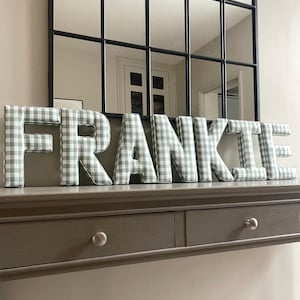 May include: The name "FRANKIE" is spelled out in large, three-dimensional letters. Each letter is covered in a light green and white gingham fabric. The letters are displayed on a gray console table, with a large black-framed mirror in the background.