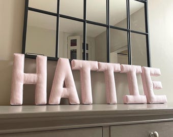 May include: A set of six freestanding fabric letters spelling the name HATTIE. Each letter is constructed from a light pink and white pinstriped fabric with visible stitched seams along the edges, creating a soft, padded, three-dimensional appearance.