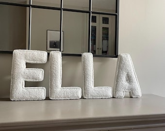 May include: Four freestanding, plush, white, textured fabric letters spelling the name ELLA. Each letter has a soft, fuzzy surface and a thick, padded construction, standing upright in a row to form the name ELLA in capital block letters.
