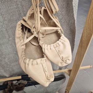 May include: A pair of handmade, light beige leather shoes with intricate stitching and tie-up laces. The shoes are suspended by their laces against a textured gray backdrop, showcasing their rustic design and craftsmanship.