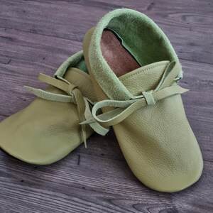 May include: A pair of soft, light green leather moccasin-style baby shoes. The shoes have a rounded toe, a light green suede collar, and a leather tie closure. The shoes are designed for infants and toddlers.