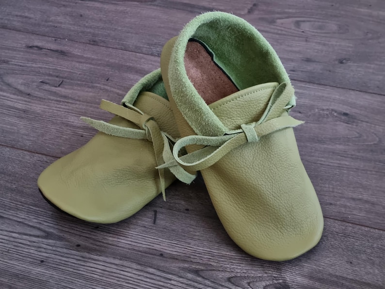 May include: A pair of soft, light green leather moccasin-style baby shoes. The shoes have a rounded toe, a light green suede collar, and a leather tie closure. The shoes are designed for infants and toddlers.