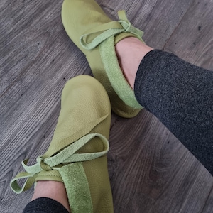 May include: A pair of light green leather moccasin-style slippers. The slippers have a rounded toe and a suede-like band around the ankle, with a matching tie. The slippers are shown on a wooden floor.