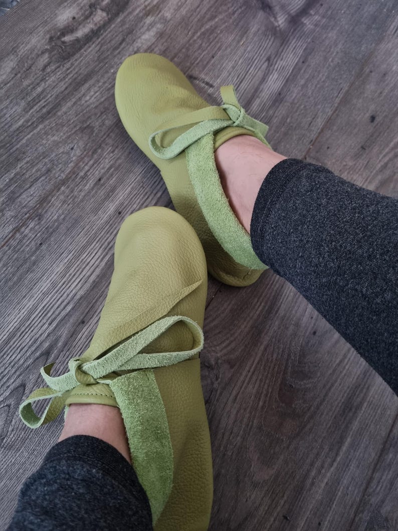 May include: A pair of light green leather moccasin-style slippers. The slippers have a rounded toe and a suede-like band around the ankle, with a matching tie. The slippers are shown on a wooden floor.