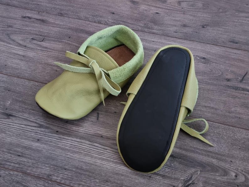 May include: A pair of soft-soled baby moccasins in a pale green hue. The shoes feature a light green suede collar and matching laces. The soles are black. The moccasins are designed for comfort and style.