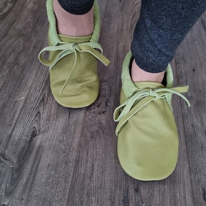 May include: A pair of soft, light green leather baby moccasins with matching laces tied in bows. The shoes have a suede-like lining and are shown on a wooden floor.