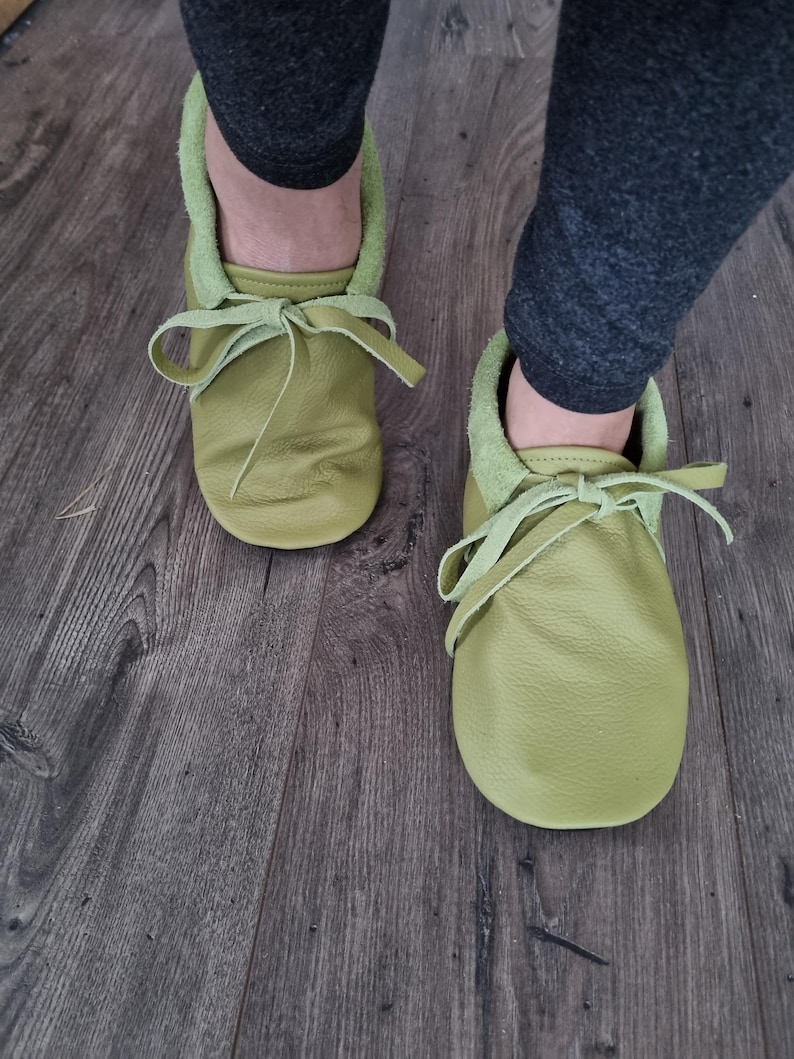May include: A pair of soft, light green leather baby moccasins with matching laces tied in bows. The shoes have a suede-like lining and are shown on a wooden floor.