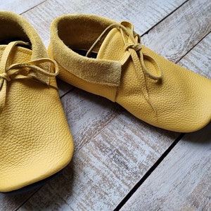 May include: A pair of yellow leather moccasins with laces tied in a bow. The moccasins are on a light wood background.