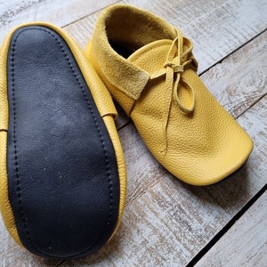May include: A pair of yellow leather moccasins with black leather soles. The moccasins have laces and are shown on a wooden surface.