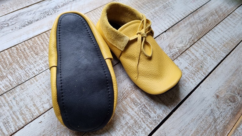 May include: A pair of yellow leather moccasins with black leather soles. The moccasins have laces and are shown on a wooden surface.