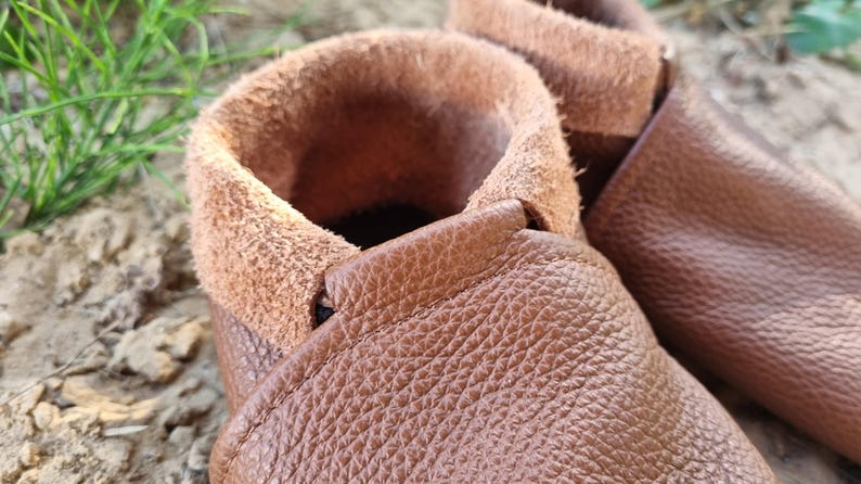 May include: Close-up of a pair of brown leather moccasins with a soft, fuzzy lining. The moccasins have a simple design and are made of high-quality leather.