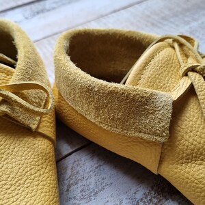 May include: A pair of yellow leather moccasins with laces tied in a bow. The moccasins have a suede lining and a textured leather exterior.