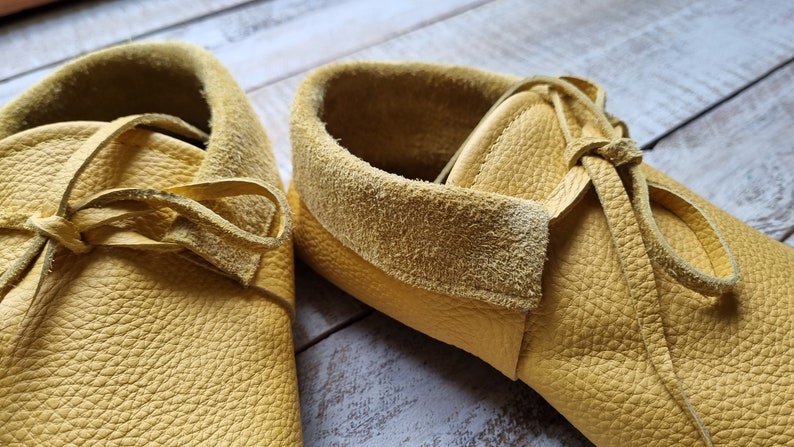 May include: A pair of yellow leather moccasins with laces tied in a bow. The moccasins have a suede lining and a textured leather exterior.