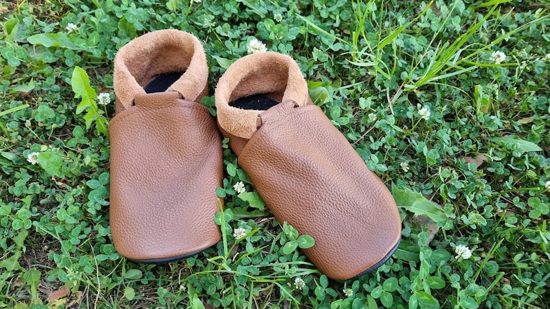 May include: A pair of brown leather moccasins with a suede lining. The moccasins are sitting on a bed of green grass.