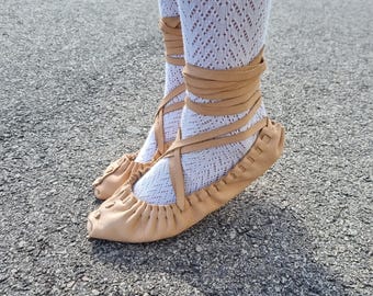May include: A pair of light tan leather ballet flats with criss-cross ankle ties. The shoes are worn with white patterned tights. The image is taken outdoors on a paved surface.