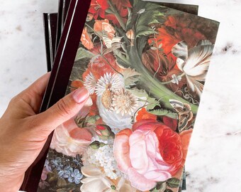 May include: A stack of three journals with a burgundy cover and a floral design on the front. The design features a variety of flowers, including roses, lilies, and daisies, in shades of pink, red, white, and green.