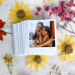 May include: A Polaroid photo of a woman and a young girl smiling at the camera. The woman is wearing a light blue and white tie-dye shirt and the girl is wearing a white tank top. They are both holding a pink flower.