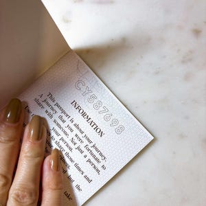 Op de afbeelding: Open paspoortpagina met de tekst "INFORMATION" en het serienummer "CY587698". De tekst beschrijft een reis die je met iemand moet delen. De pagina wordt vastgehouden door een hand met bruine nagellak.