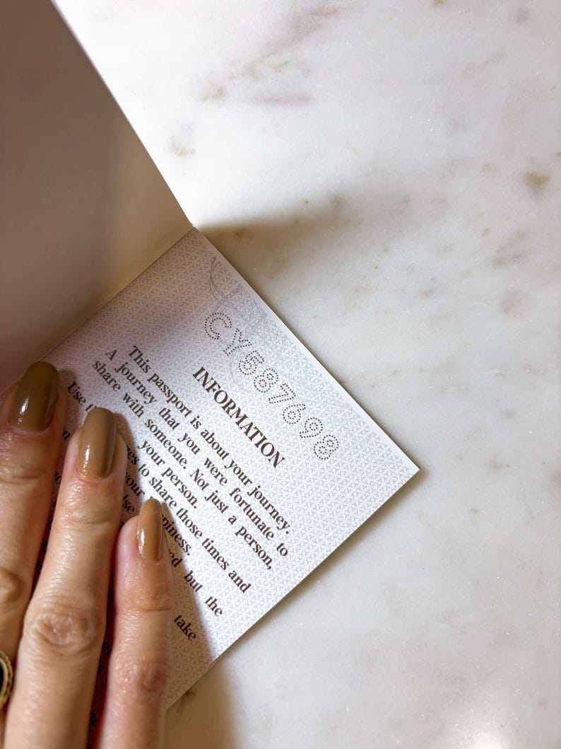Op de afbeelding: Open paspoortpagina met de tekst "INFORMATION" en het serienummer "CY587698". De tekst beschrijft een reis die je met iemand moet delen. De pagina wordt vastgehouden door een hand met bruine nagellak.