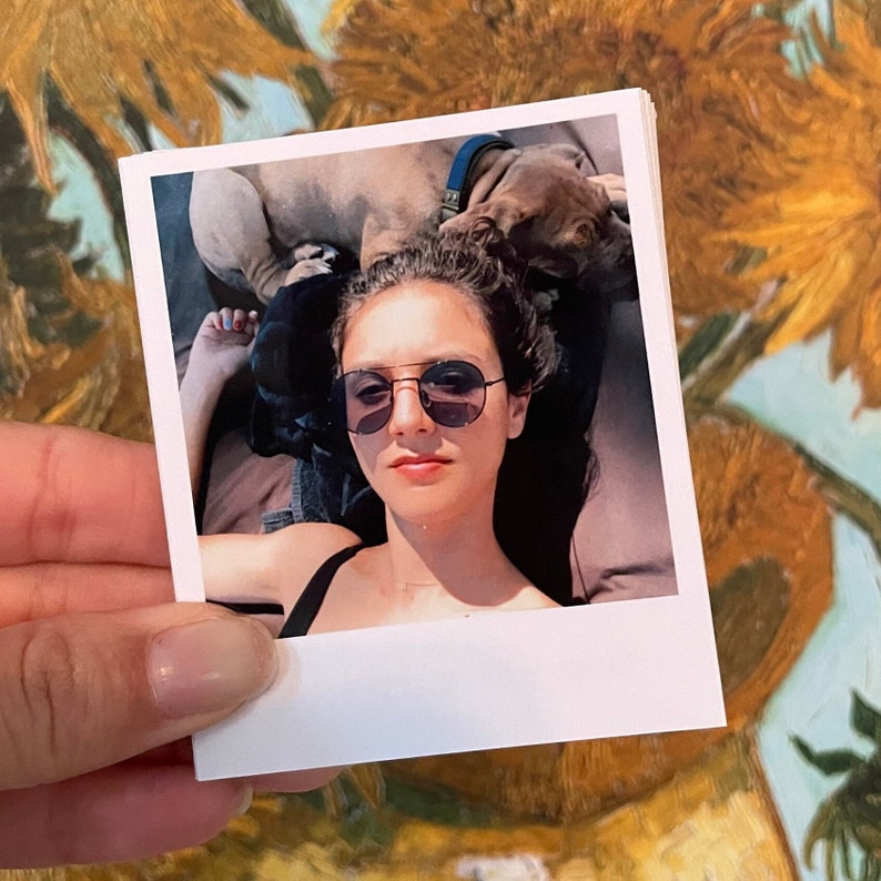 May include: A woman wearing sunglasses and a black tank top is lying down with a brown dog on top of her. The photo is printed on a white Polaroid-style photo paper.